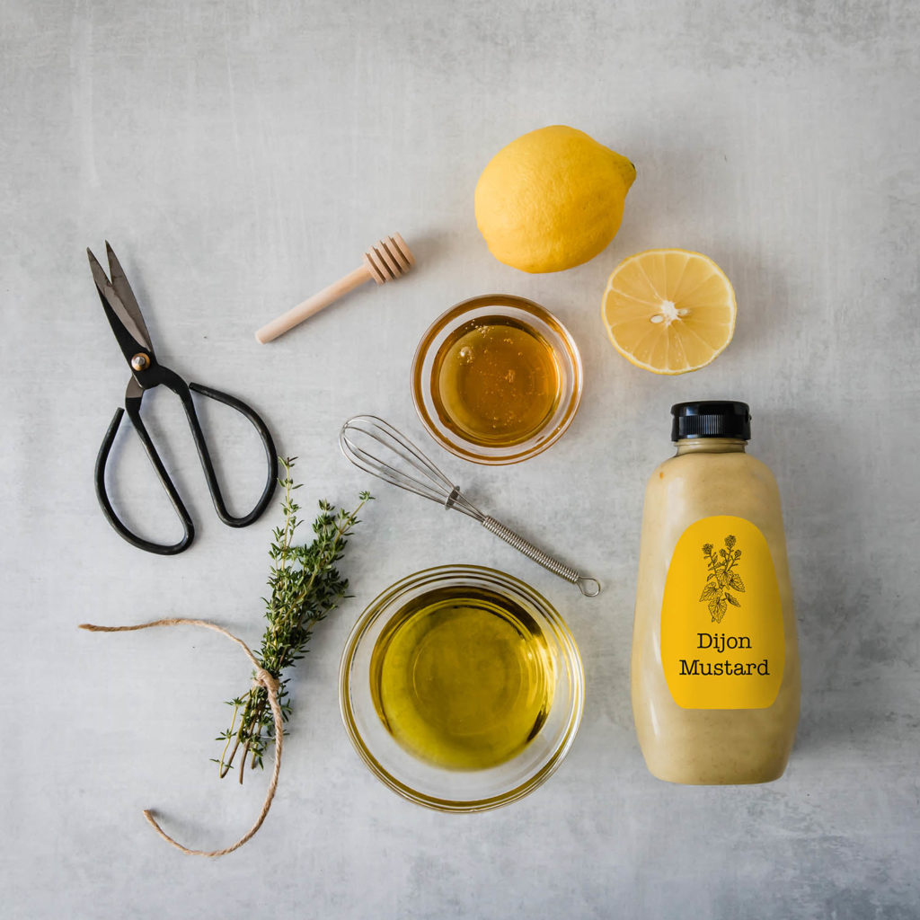 An ingredient flat lay photograph with lemons, thyme, honey, dijon mustard and olive oil, shot from above.